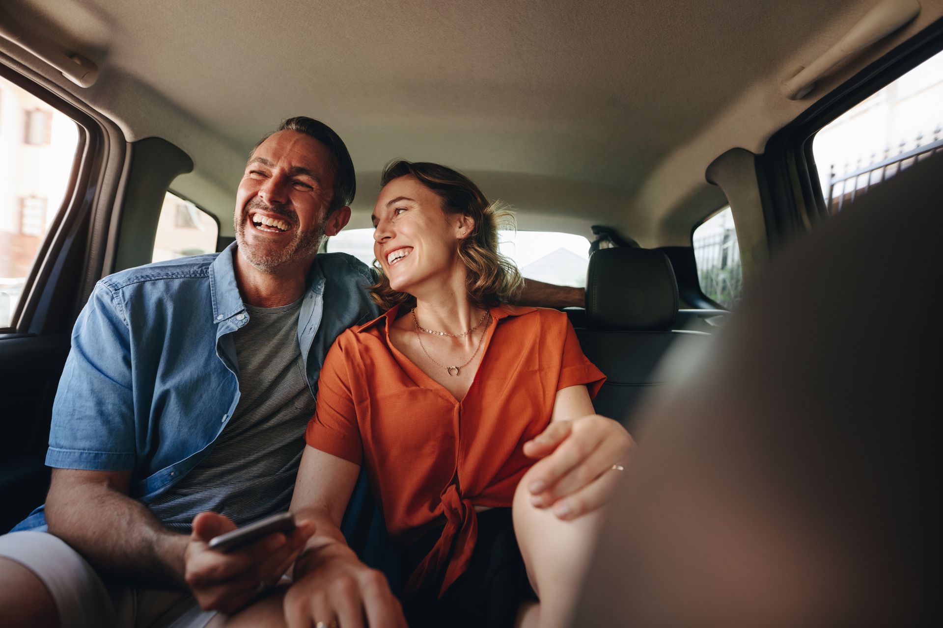 Couple souriant en voiture, l'homme tenant un téléphone, la femme portant un haut orange, lumière naturelle, expressions joyeuses.