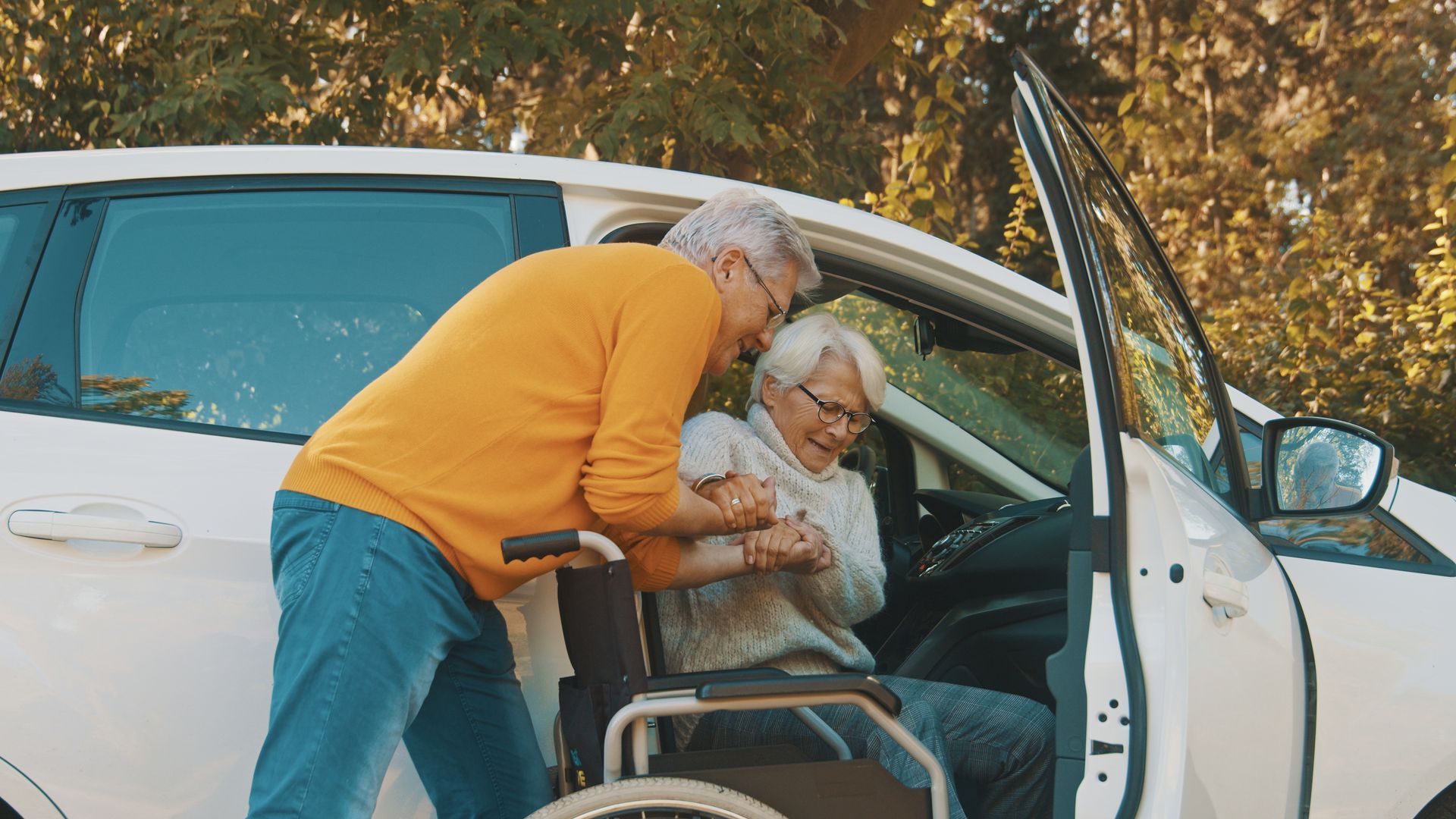 Un homme aide une femme à monter dans une voiture. Voiture blanche, homme en chemise orange, femme en fauteuil roulant. Lumière du soleil.