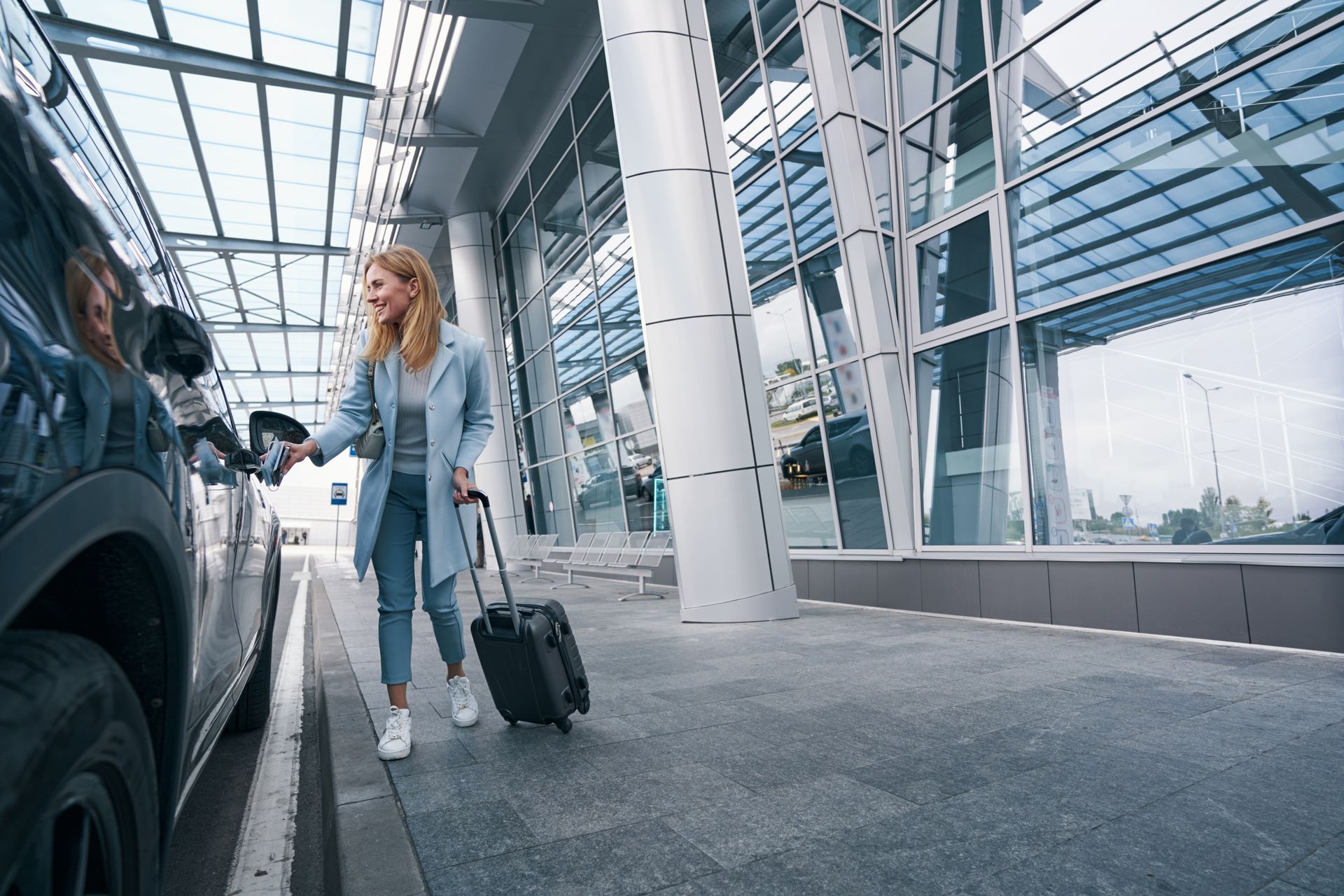 Une femme sort d'un SUV noir à l'aéroport, souriante et portant des bagages.