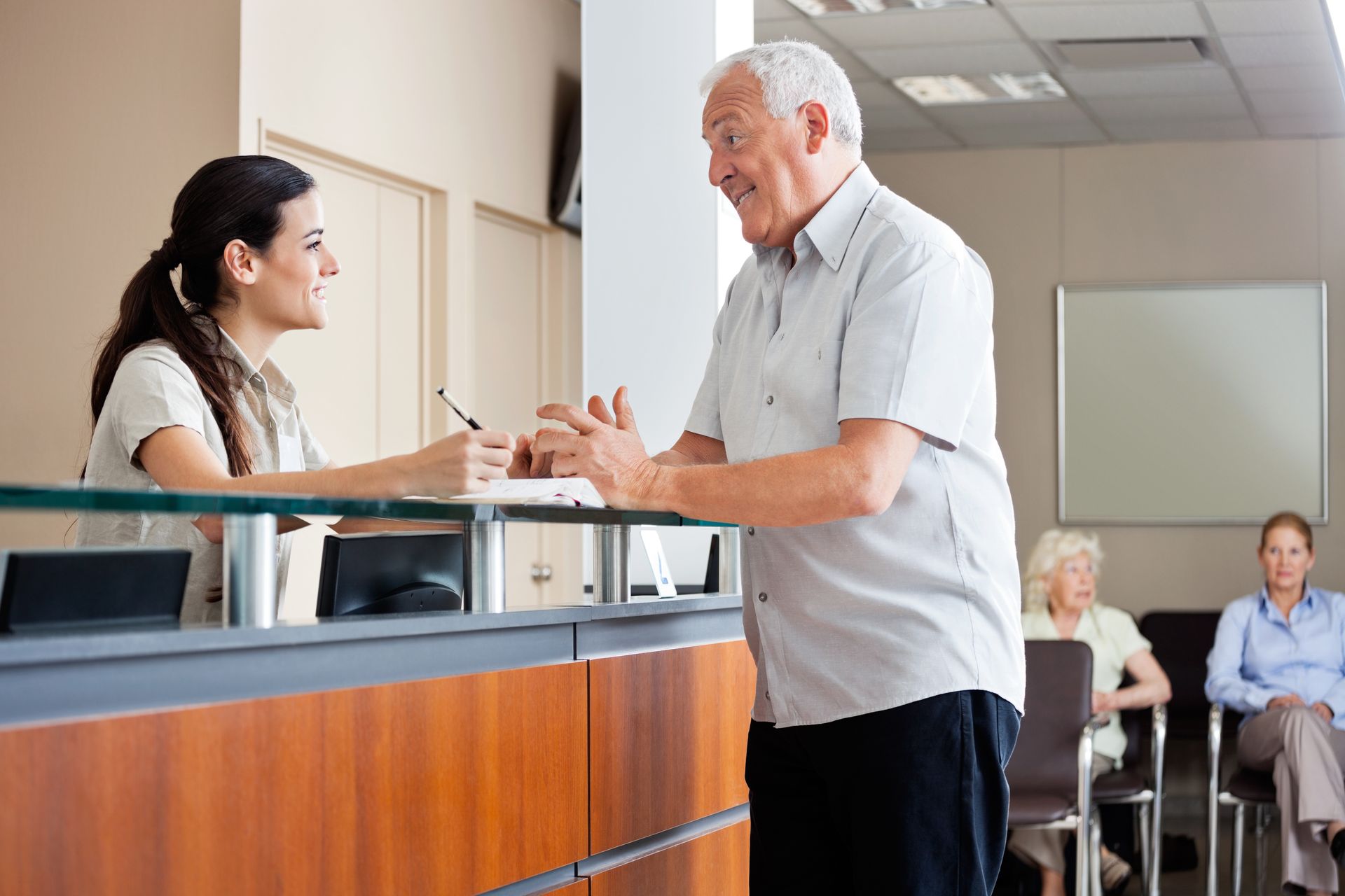 Un homme parle à une réceptionniste à un bureau ; d'autres patients attendent en arrière-plan.