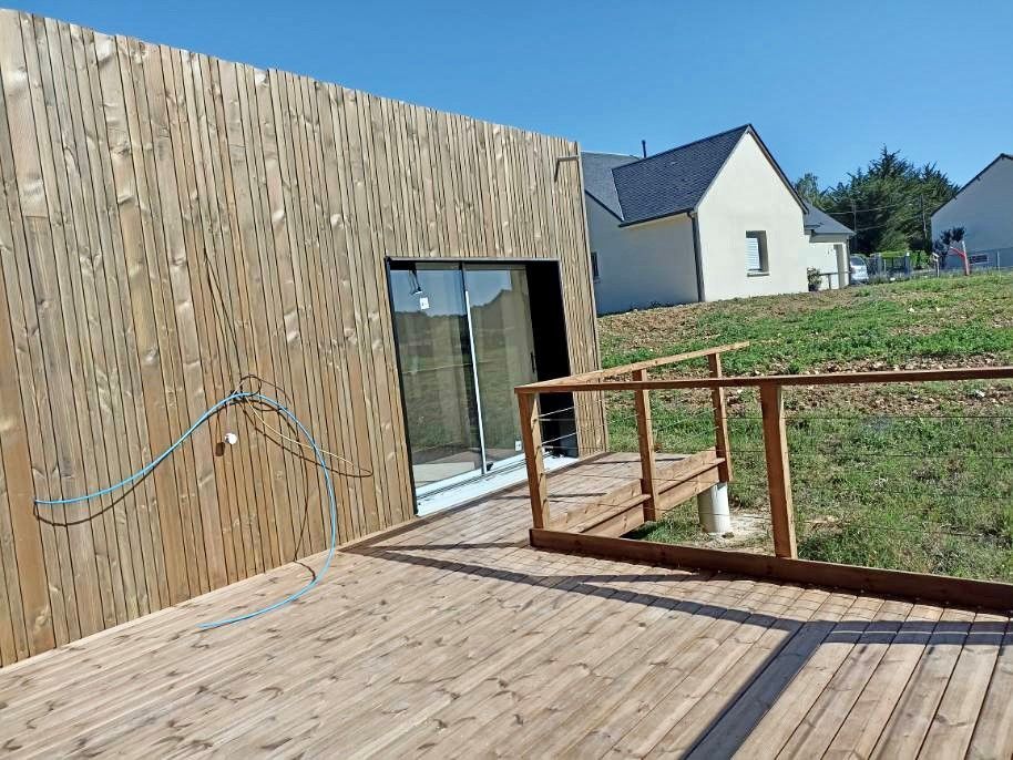 Terrasse en bois avec rampe menant à un bâtiment moderne avec un mur lambrissé de bois, situé dans une cour herbeuse.