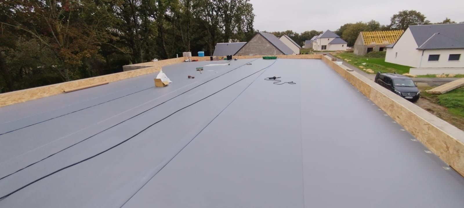 Vue de dessus d'un toit plat en construction ; surface grise, bords en bois, arbres et maisons environnants.