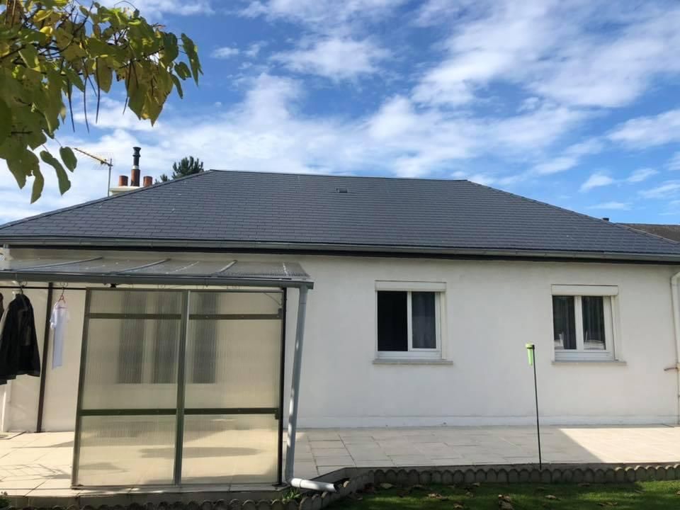 Maison blanche au toit gris foncé. Une petite véranda se trouve à gauche et deux fenêtres sont visibles.