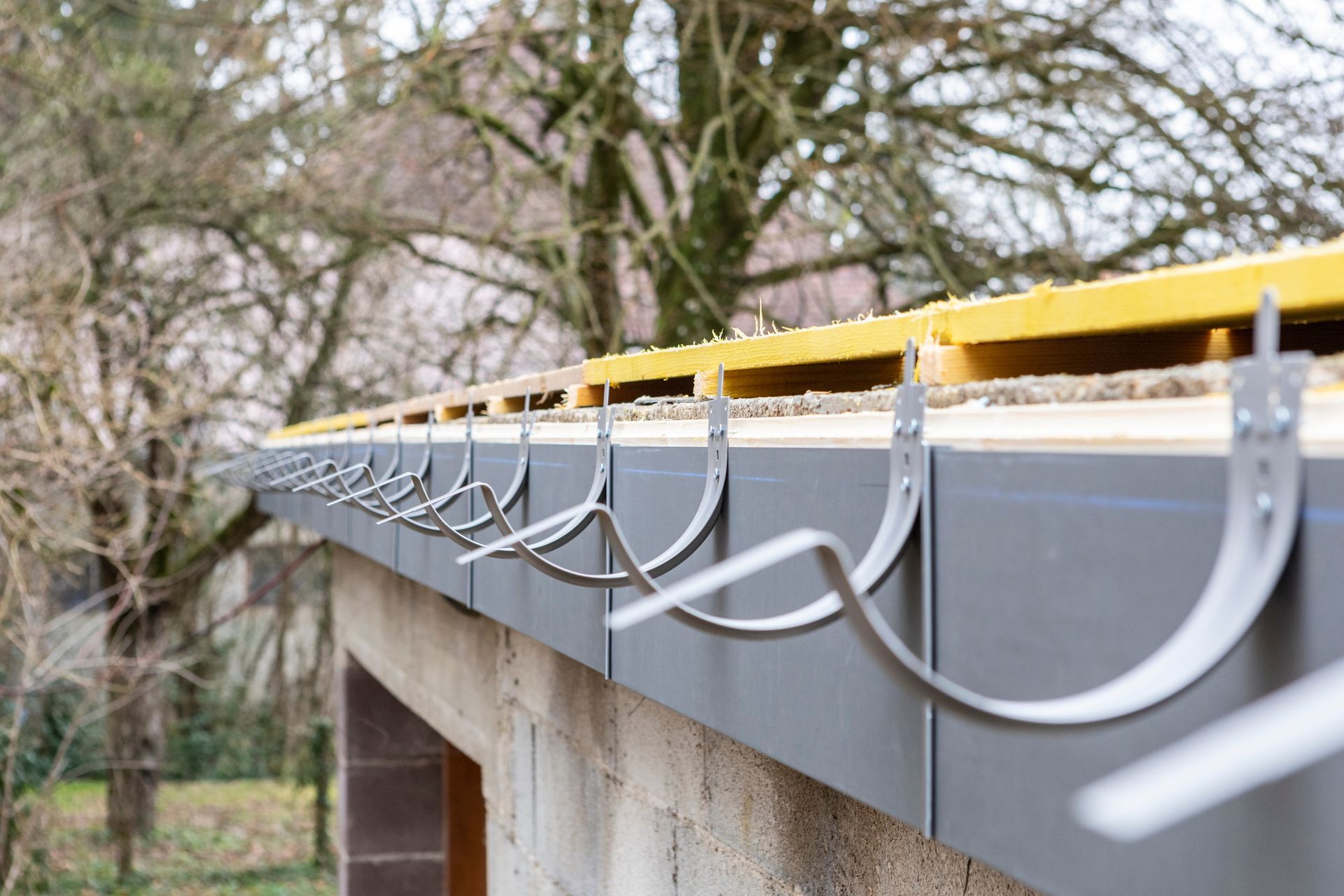 Gouttière grise avec des supports métalliques fixés au bord d'un toit, sur fond d'arbre flou.