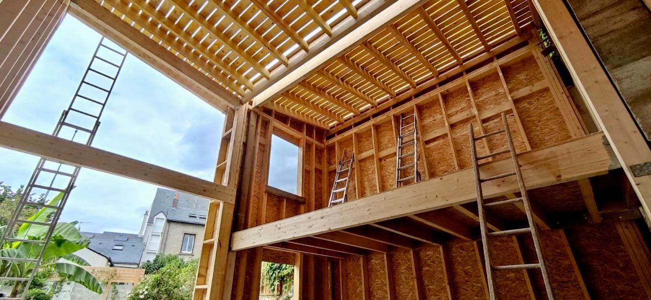 Vue intérieure d'une maison en bois en construction ; une échelle s'appuie contre le mur.