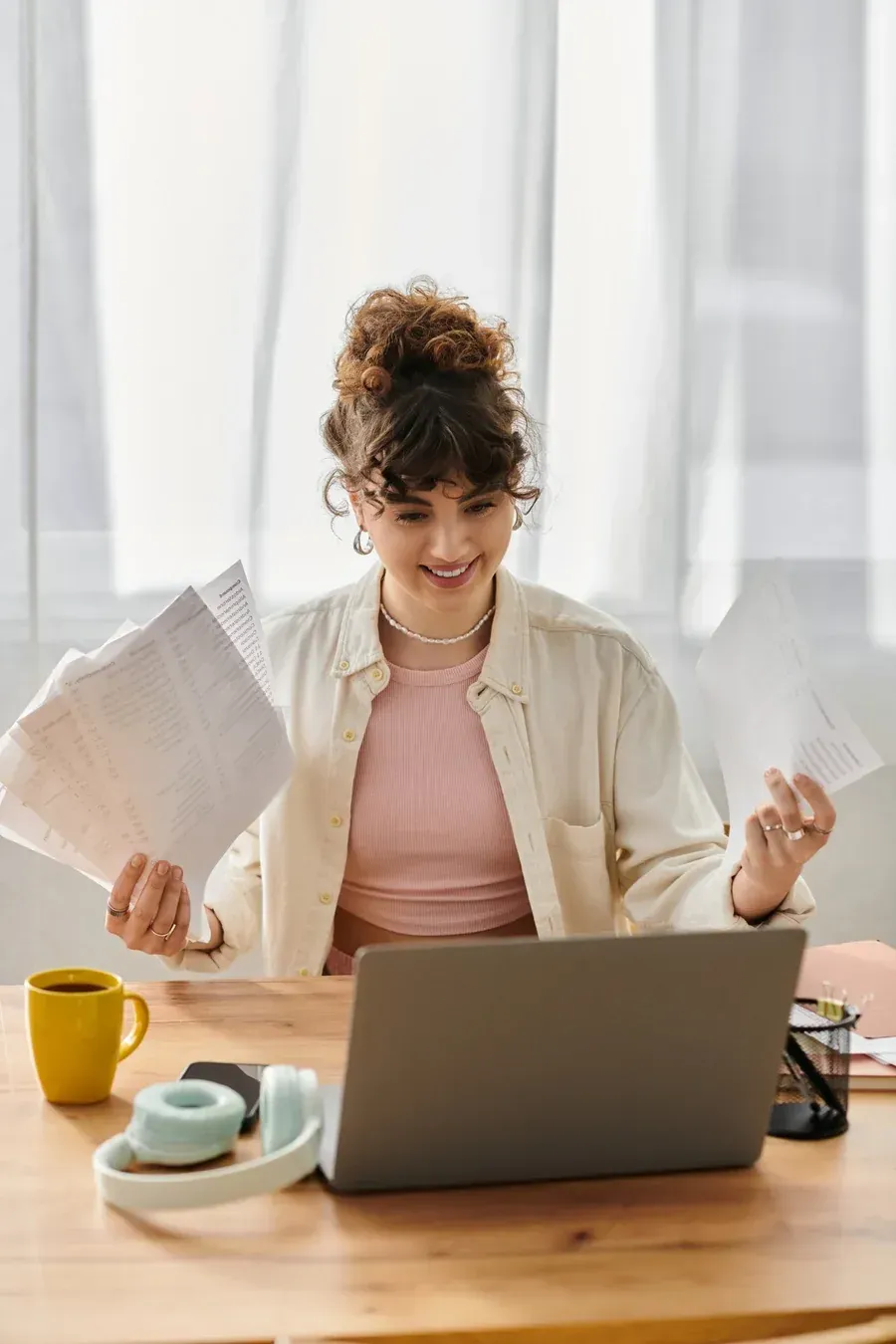 Mujer sentada con papeles en la mano, sonriendo a su portátil. Taza de café, auriculares y bloc de notas visibles.