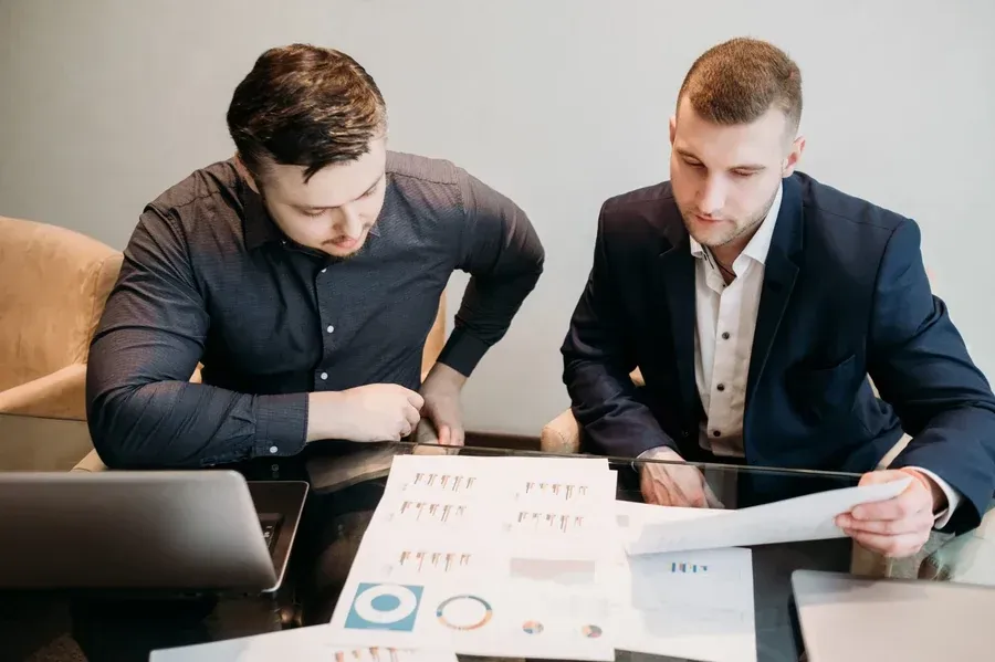 Dos hombres revisando documentos en una mesa; uno con traje y otro con camisa abotonada.