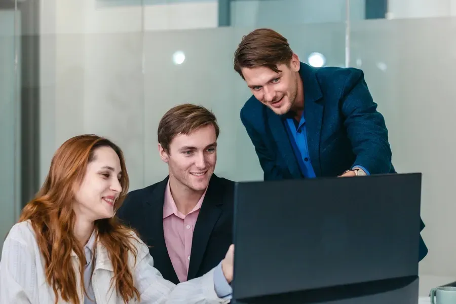 Tres hombres de negocios mirando una pantalla de computadora, uno de ellos señalando, sonriendo, en una oficina luminosa.