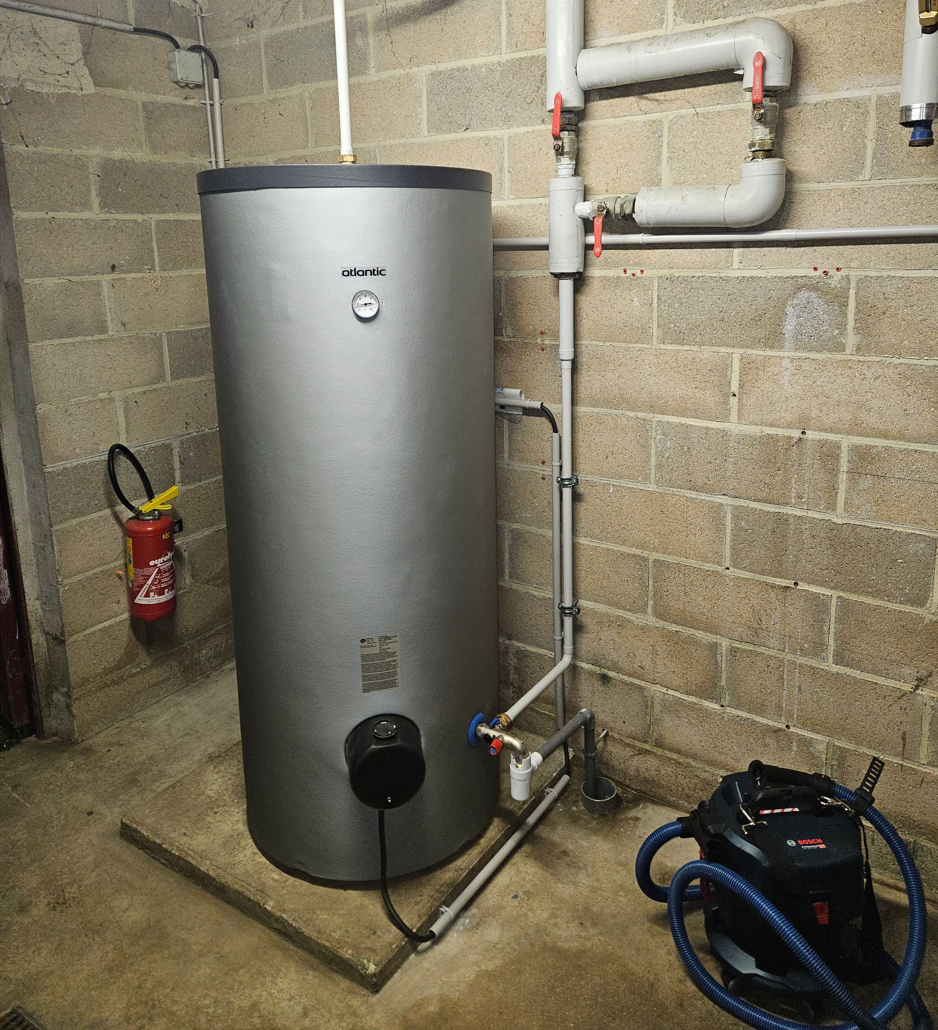 Un chauffe-eau cylindrique argenté est installé dans un sous-sol en parpaings, avec des tuyaux, des vannes, un extincteur et d'autres équipements.