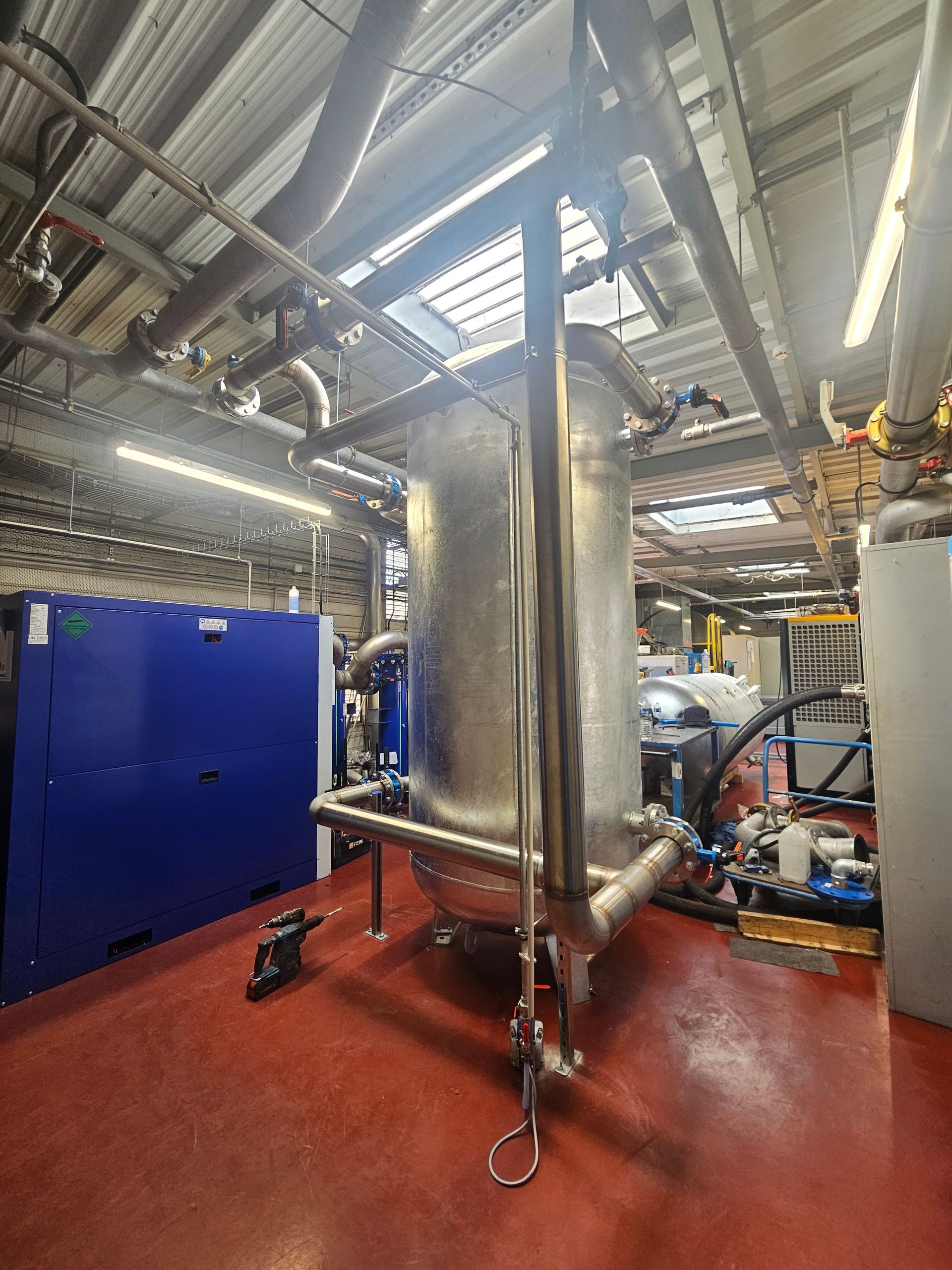 Une grande cuve industrielle métallique sous pression se dresse dans une installation aux sols rouges, avec des tuyaux et des machines bleues à proximité.