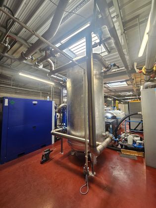 Une grande cuve industrielle métallique sous pression se dresse dans une installation aux sols rouges, avec des tuyaux et des machines bleues à proximité.