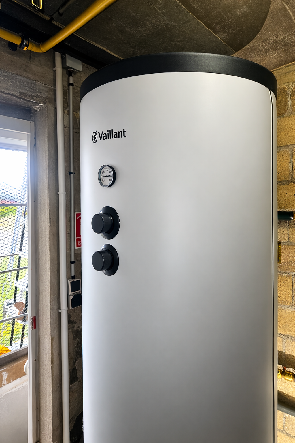Un grand réservoir d'eau chaude Vaillant cylindrique gris clair, équipé d'un thermomètre et de deux cadrans, dans une buanderie.