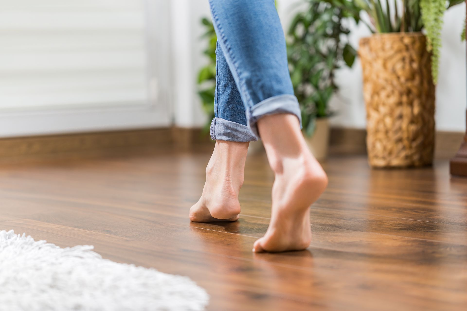 Des pieds nus marchant sur un parquet lisse en bois brun, les chevilles vêtues de denim visibles dans un intérieur moderne et lumineux.