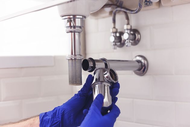 Des mains gantées de gants en nitrile bleus fixent un siphon chromé en forme de P à un tuyau d'évacuation de lavabo contre un mur carrelé blanc.