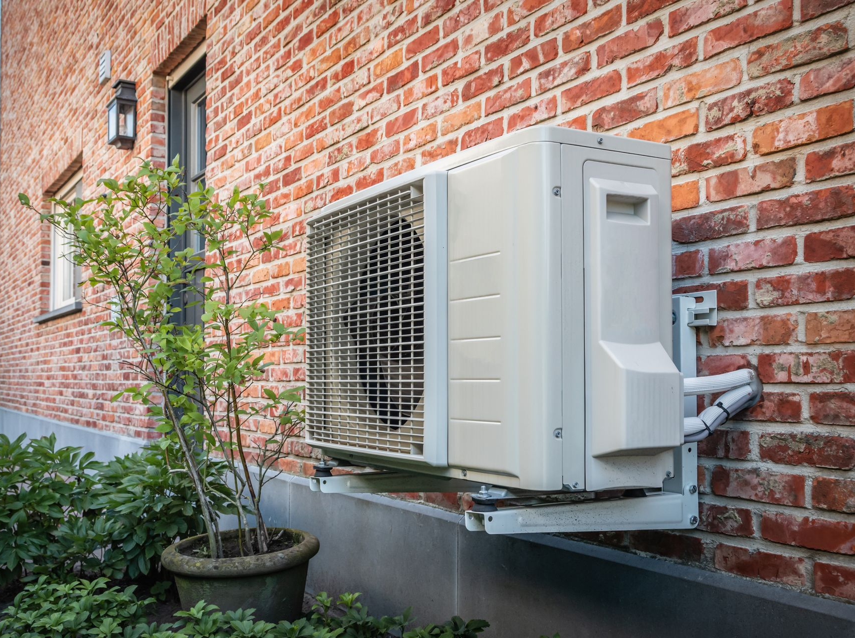 Un climatiseur extérieur blanc, fixé sur un mur de briques rouges, à côté d'une petite plante en pot et d'une porte sombre.