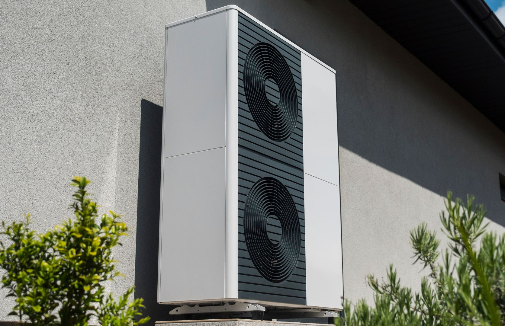 Une grande unité de pompe à chaleur extérieure, blanche et gris foncé, montée sur un mur extérieur clair à côté de buissons verts.