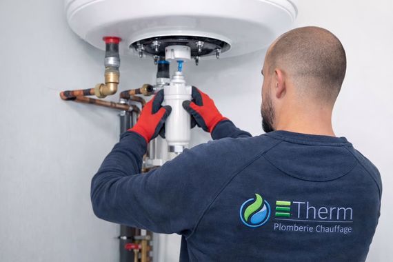 Un plombier portant des gants rouges travaille sur un chauffe-eau blanc raccordé à des tuyaux en cuivre.