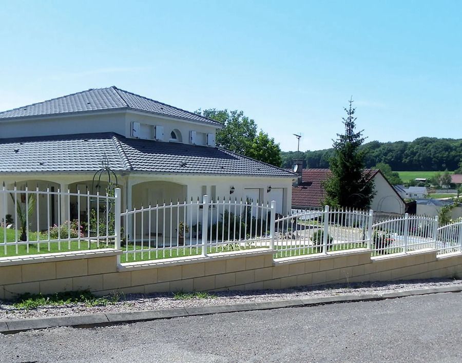 Maison blanche derrière une clôture en métal blanc sur un muret de pierre ; arbres et ciel bleu en arrière-plan.