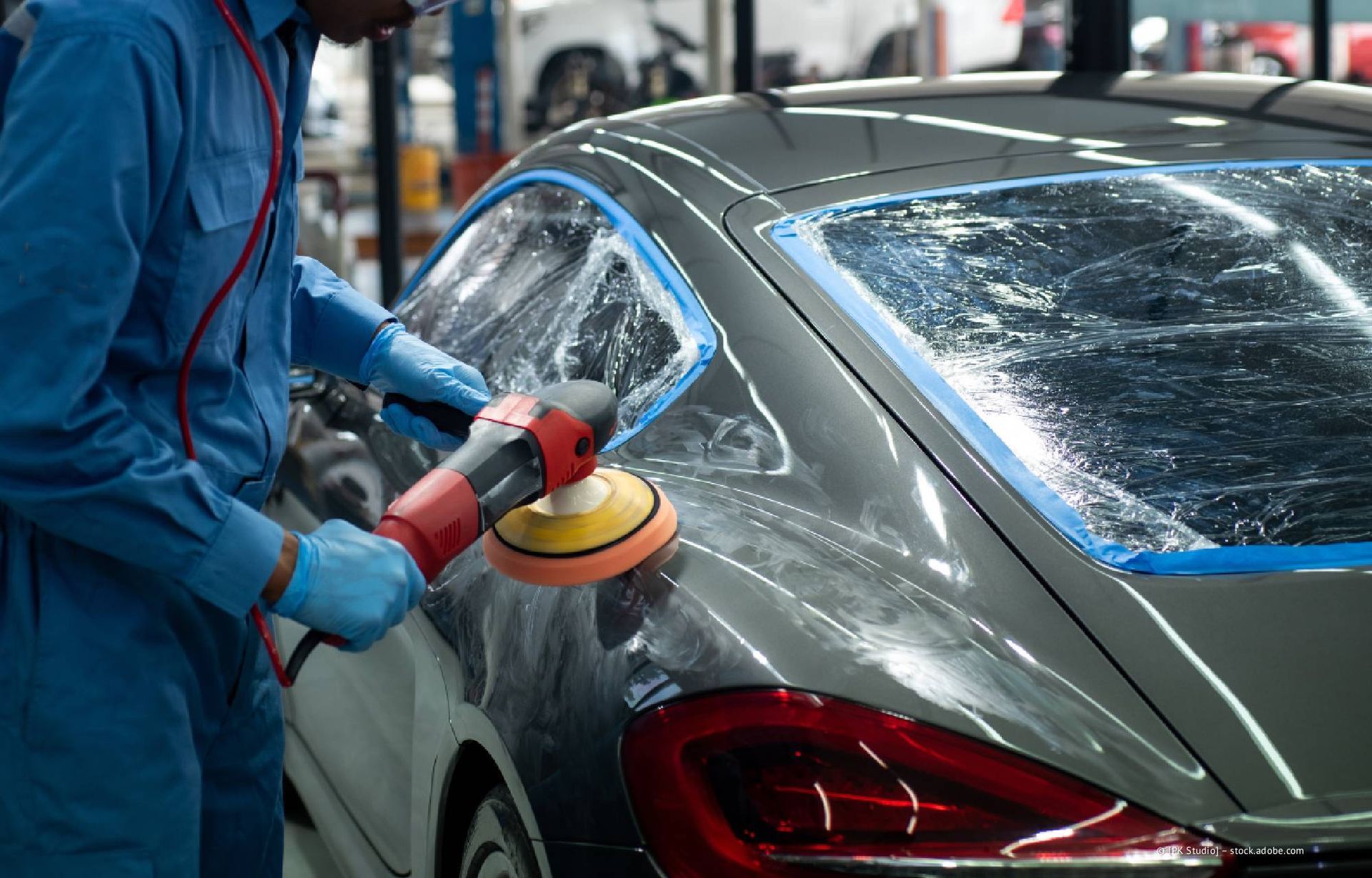 Eine Person in blauem Overall poliert ein graues Auto mit einer Poliermaschine.