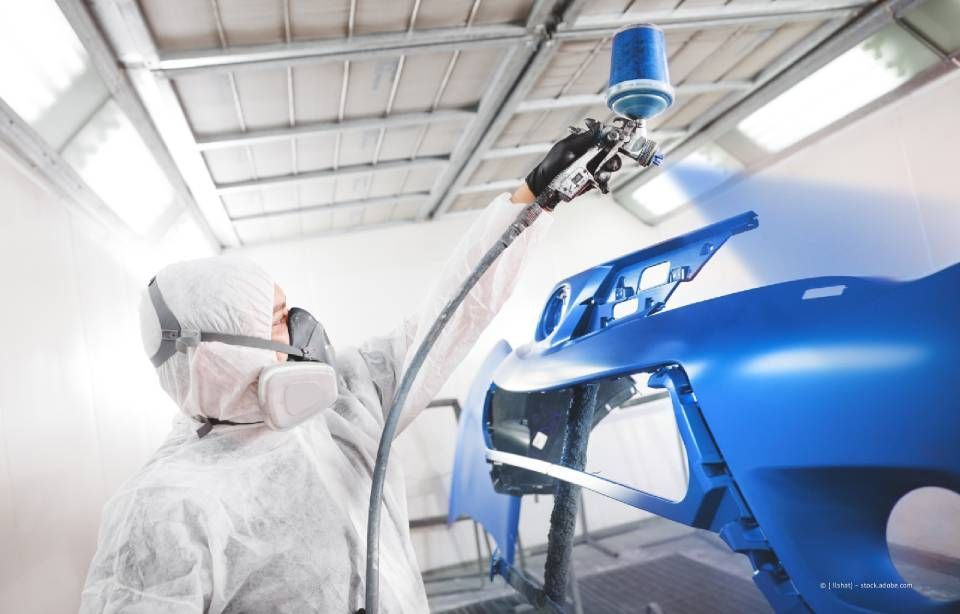 Eine Person in Schutzkleidung besprüht in einer Lackierkabine eine Autostoßstange mit blauer Farbe.