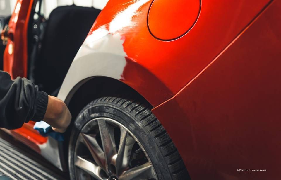 Eine Person, die mit einer Spritzpistole Farbe auf die Seitenwand eines Autos sprüht und dabei eine Atemschutzmaske trägt.