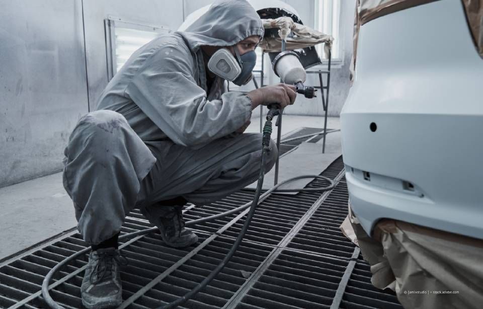 Eine Person in Schutzkleidung lackiert ein blaues Autoteil in einer Lackierkabine.