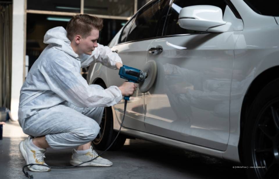 Eine Person in einem weißen Anzug benutzt in einer Autowerkstatt eine Heißluftpistole und ein Schleifgerät, um eine Autotür zu lackieren.