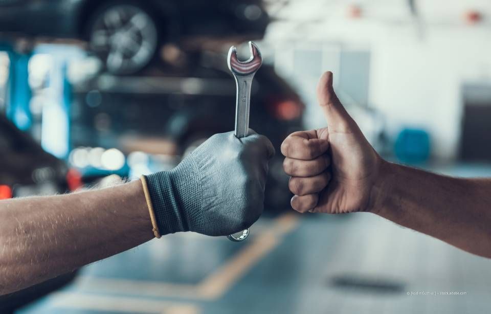 Ein Daumen hoch über dem Automotor, ein Mechaniker hält einen Schraubenschlüssel in der Hand, beide sind sich einig.