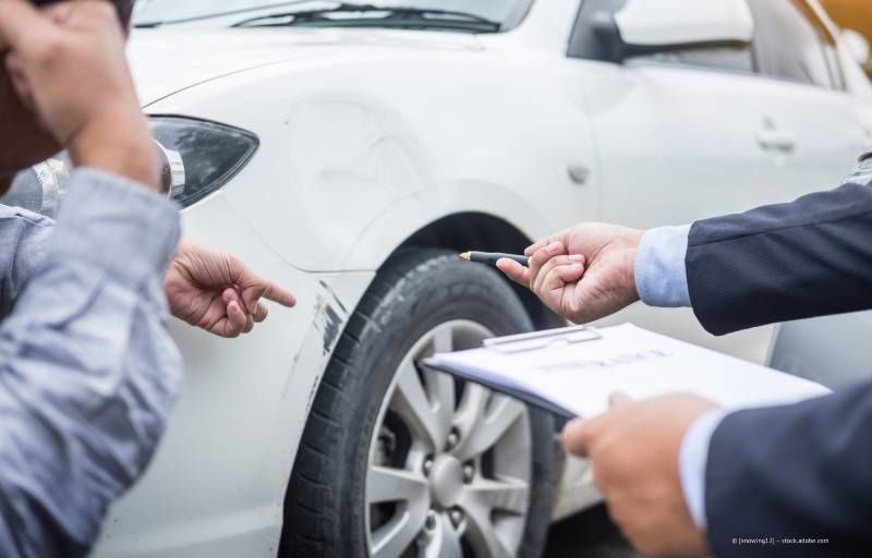 Zwei Männer begutachten den Schaden an der Seite eines weißen Autos, möglicherweise nach einem Zusammenstoß. Einer zeigt auf einen Kratzer.