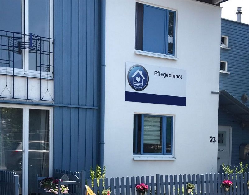 Blau-weiße Gebäudefassade mit Schild und Fenster, hinter einem kleinen Zaun.