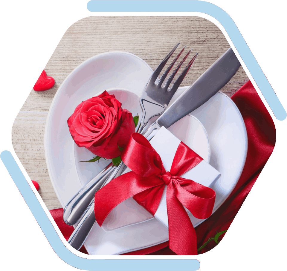 Tischdekoration zum Valentinstag: herzförmige Teller, rote Rose, Geschenkbox mit roter Schleife, Silberbesteck und roter Satin.