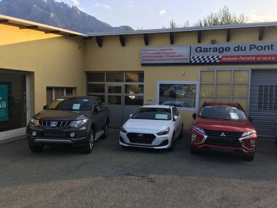 Trois voitures garées devant le Garage du Pont, un garage jaune avec des montagnes en arrière-plan.