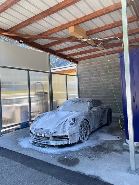 Voiture de sport recouverte de mousse, garée sous un abri, en cours de lavage ; de l'eau se répand sur le sol en béton.