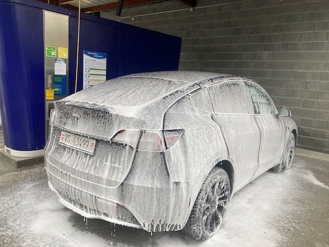 Voiture blanche recouverte de mousse dans une station de lavage, garée à côté d'une structure bleue.