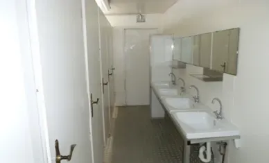 Öffentliche Toilette mit mehreren Waschbecken und Spiegeln; die Kabinen befinden sich an einer Wand.