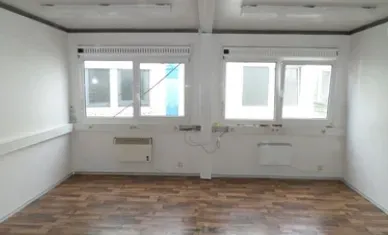 Leeres Bürointerieur mit zwei Fenstern, braunem Boden, weißen Wänden und zwei Heizkörpern.