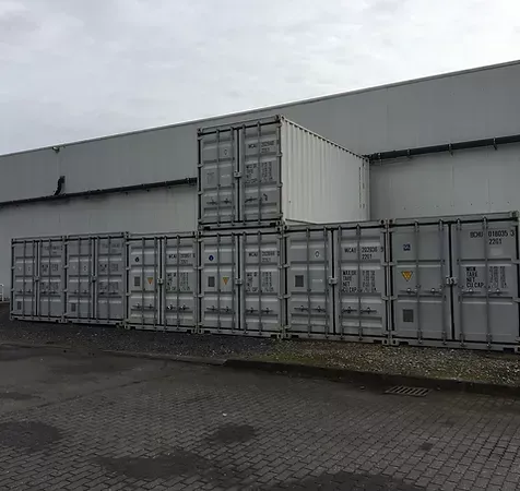 Vor einem weißen Gebäude auf grauem Asphalt gestapelte Schiffscontainer.