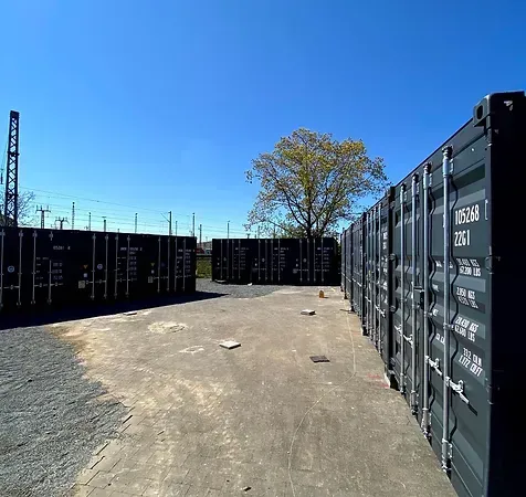 Reihen schwarzer Lagercontainer auf einem Freigelände an einem sonnigen Tag. Ein Baum und Bahngleise sind zu sehen.