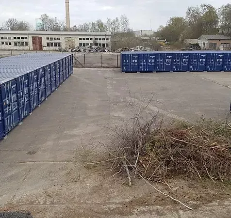 Reihen blauer Schiffscontainer auf einem Betonplatz, im Hintergrund Gebäude und ein Schornstein.