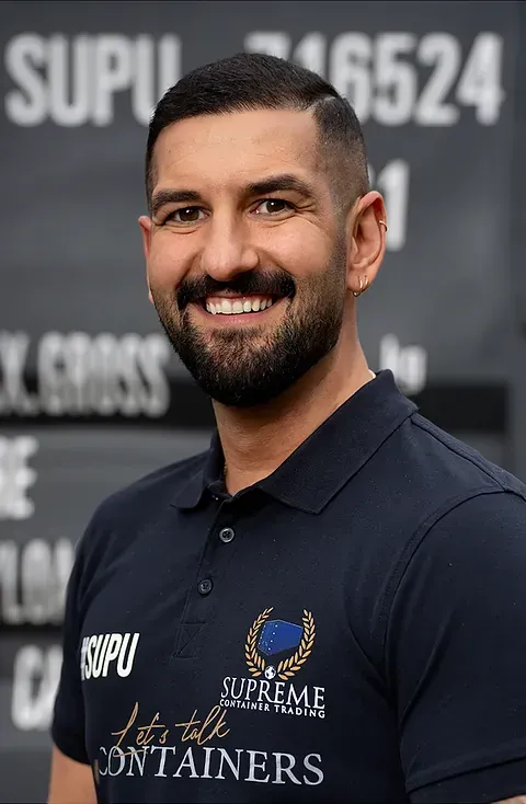 Ein Mann mit dunklen Haaren und Bart lächelt. Er trägt ein dunkelblaues Polohemd mit Logo und steht vor einem Container.