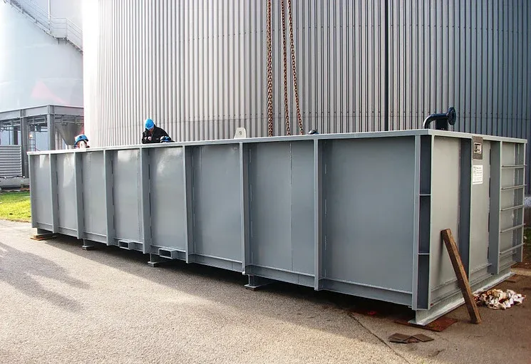 Ein grauer Industrietank steht im Freien, daneben ein großer Metallzylinder. Zwei Arbeiter sind obenauf zu sehen.