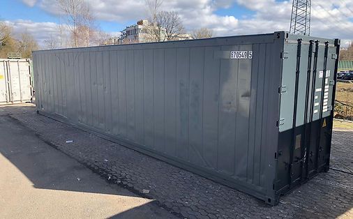 Ein dunkelgrauer Schiffscontainer steht im Freien auf Asphalt, neben einem weißen Container und Bäumen.