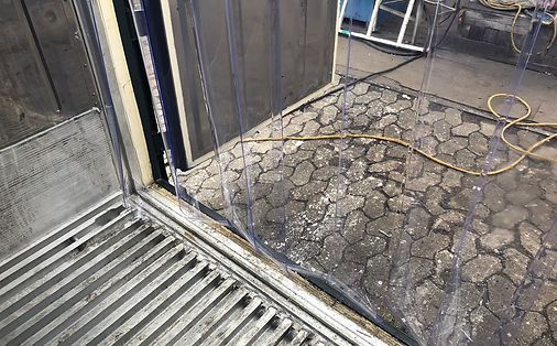 Im Türrahmen hängen durchsichtige Plastikstreifen, die den grau-schwarzen, rissigen Gehweg verdecken.