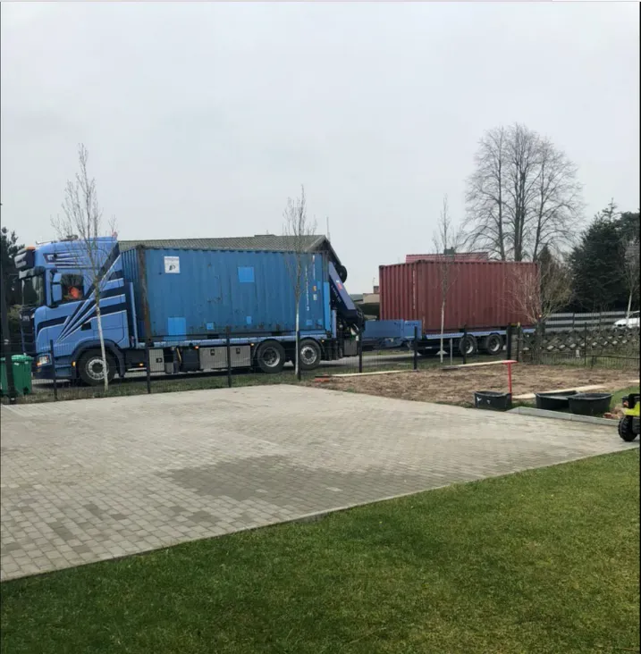 Blaue und rote Schiffscontainer auf Lastwagen, die auf dem Gehweg neben grünen Wiesen und Bäumen parken.