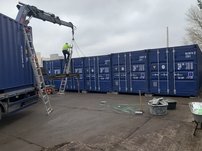 Ein Arbeiter bewegt mit einem Kran Frachtcontainer. Blaue Container und ein Lkw an einem bewölkten Tag.