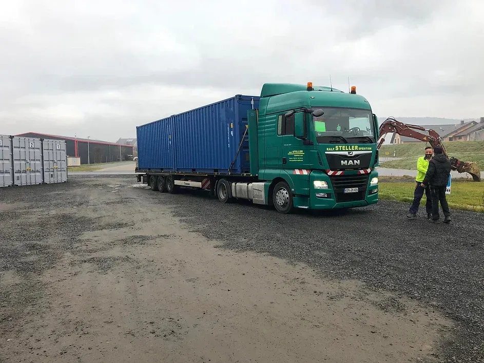 Grüner Sattelzug mit blauem Containeranhänger; zwei Personen rechts; Betonblöcke im linken Hintergrund; bewölkter Tag.