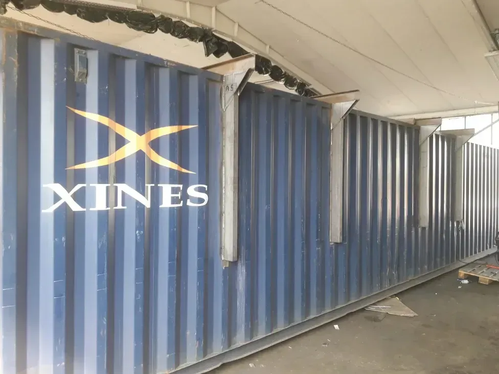 Blauer Schiffscontainer mit der gelben Aufschrift „XINES“ und Metallstützen.