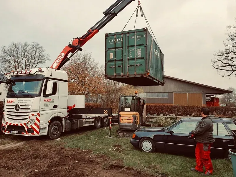 Ein LKW-Kran hebt einen grünen Schiffscontainer an; eine Person beobachtet das Geschehen