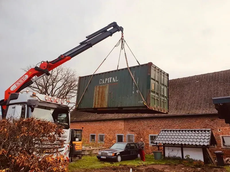 Ein Kran hebt einen grünen, mit „CAPITAL“ beschrifteten Schiffscontainer in der Nähe eines Backsteingebäudes an