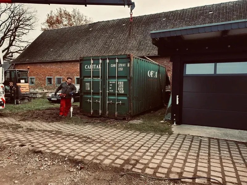 Ein grüner Schiffscontainer wird mit einem Kran in der Nähe einer Scheune und einer Garage platziert. Männer sind anwesend.